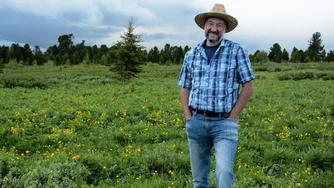 Man farmer in a straw hat on a background of a high-mountain field with flowers Stock Footage 83034206