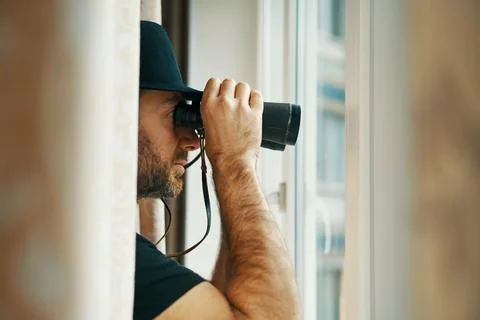 Man in fedora hat looking through binoculars spying from window Stock Photos