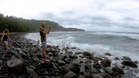 Man films ocean waves while woman steps onto stony beach, Pololū Valley Hawaii Stock Footage 98426326