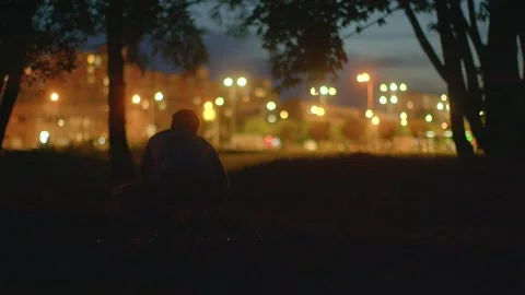 A man finds solace under a tree in a tranquil night park Видео 334032186