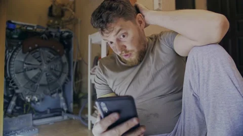 A man fixes a washing machine and watches instructions on his phone Stock Footage 155336742