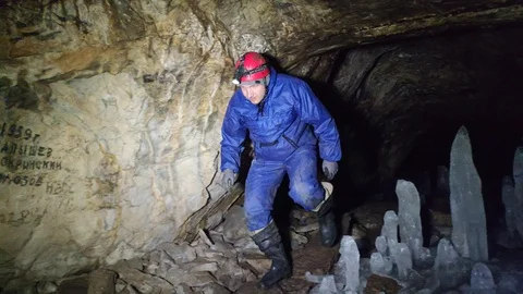 A man with a flashlight walks through a cave past stalagmites and stalactides Stock Footage 129495099
