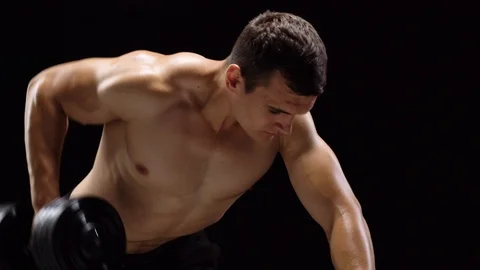 Man flexes his hands with dumbbells, training his back on a black background in Stock Footage 95033562