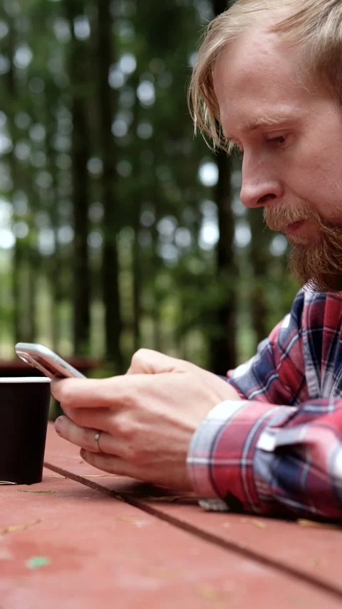 Man focused on smartphone while sitting at wooden table in serene forest setting Stock-Footage 322122777