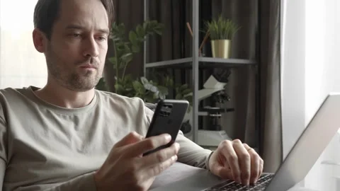 The man is focused on using a smartphone and laptop for remote work at home Stock Footage 253403210