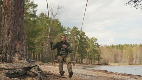Man in the forest swing on a swing drinking coffee. Rope swing on a forest lake. 스톡 동영상 197286629
