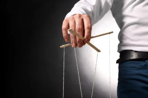 Man in formal outfit pulling strings of puppet on dark background, closeup... Stock Photos