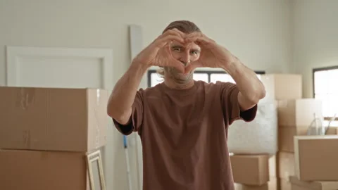 Man forms heart shape with hands amid packed moving boxes inside a building.. Stock Footage 339195299