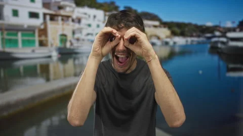 Man frames eyes with fingers and opens mouth at sunny port by moored boats;.. Video stock 314688996