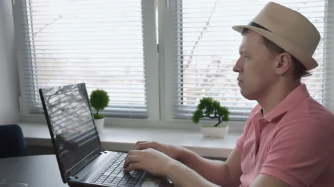 Man freelancer in hat sitting at table and typing text on laptop, window Stock Footage 155923902