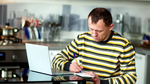 Man freelancer working at home using a laptop, making notes in a notebook Stock Footage 143160125