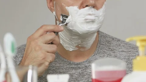 A man in front of a bathroom mirror shaves his face covered with white foam Vídeo Stock 100716725