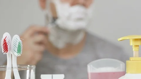 A man in front of a bathroom mirror shaves his face covered with white foam Stock Footage 100720197