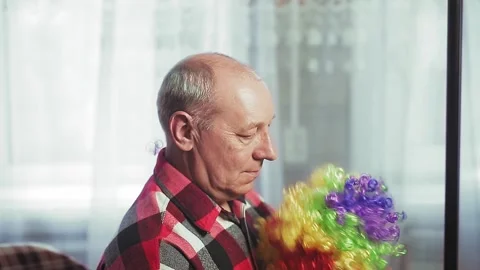 A man in front of a mirror puts on a clown wig with multi-colored curls 스톡 동영상 131751573