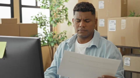 Man frowning while reading a delivery document at a computer amid stacked p.. Stock Footage 347818632