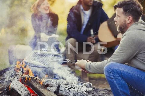 Man frying fish in grill basket over campfire near friends Stock Photos