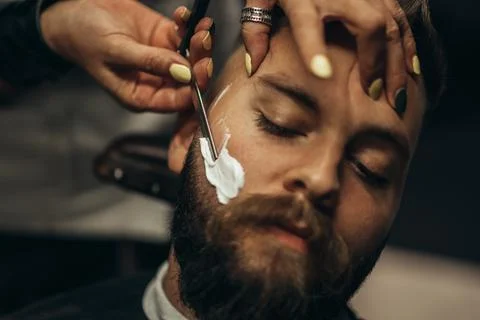 Man getting shaved with straight edge razor by hairdresser at barbershop Stock Photos