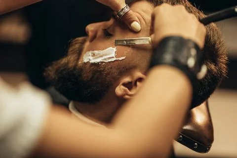 Man getting shaved with straight edge razor by hairdresser at barbershop Stock Photos