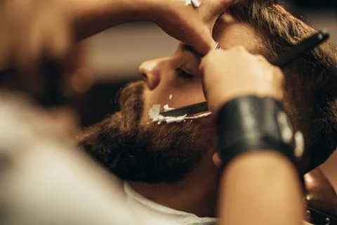 Man getting shaved with straight edge razor by hairdresser at barbershop Stock Photos