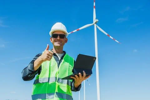 Man gives thumbs up while holding a tablet near a wind turbine on a clear s.. Stock Photos