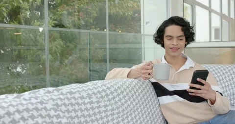 Man glancing at phone screen, scrolling and sipping coffee on couch beside glass Stock Footage 343239750