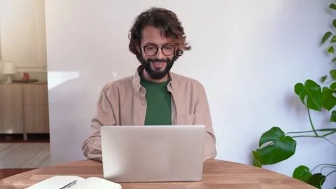 A man with glasses and a beard is sitting at a table at home using a laptop Stock Footage 264842357