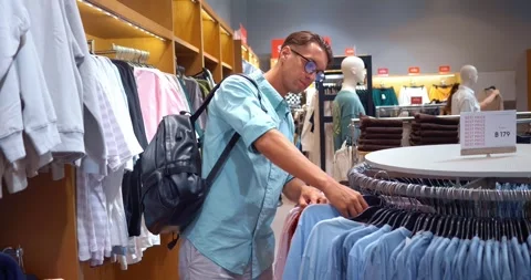 Man glasses, backpack, takes blue shirt, his choice part shopping. He likes it Stock Footage 263179086