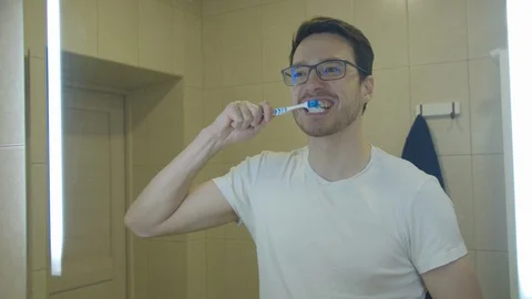 A man with glasses brushes his teeth in front of a mirror Stock Footage 128549235