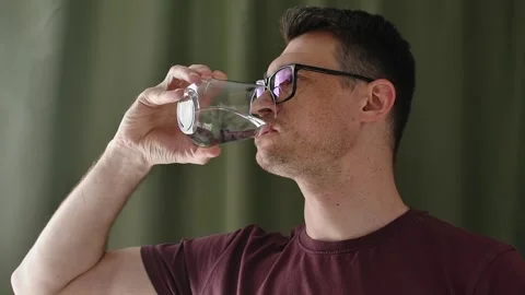 Man in glasses drinks water from clear glass at home in morning, starting simple Stock Footage 336623836