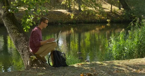 Man in Glasses With Laptop is Sitting on Tree Trunk at River Freelance Stock Footage 55547396