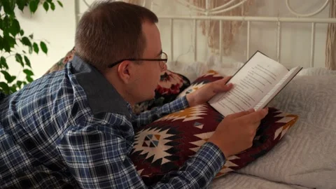 A man with glasses reading a book lying on the bed Stock Footage 157368130
