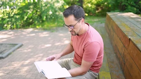 A man with glasses reads a book while sitting on a park bench on a summer day. Vídeos de archivo 247125069