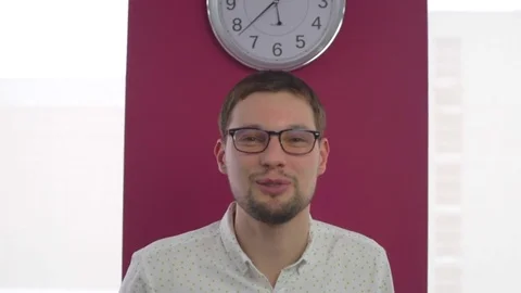 Man with glasses says something, he stands on the background with a clock. The Stock Footage 74096804