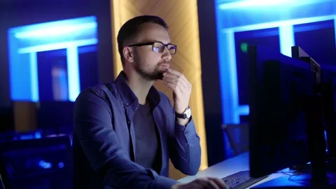 A man with glasses sits and thinks, typing on a computer in the office at night. Stockbeeldmateriaal 198057846