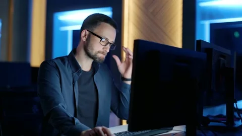 A man with glasses sits and thinks, typing on a computer in the office at night. Vídeos de archivo 198057880