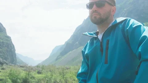 A man with glasses sits on the edge of a cliff wearing a blue anorak. Katu-Yaryk Stock Footage 166065737