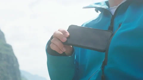 A man with glasses sits on the edge of a cliff wearing a blue anorak pulls out Stock Footage 166065803