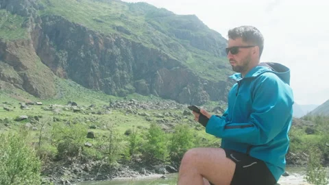 A man with glasses sits on the edge of a cliff wearing a blue anorak pulls out Stock Footage 166065880