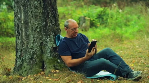 A man in glasses sits under a tree in a park and communicates in a messenger on Stock Footage 95954684