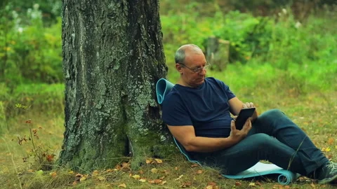 The man in glasses sits under a tree in the park and works on a tablet Stock Footage 96035730