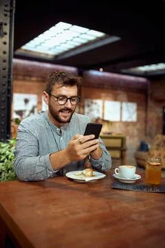 Man with glasses sitting in cafe, scrolling on his phone. Coffee break. Stock Photos