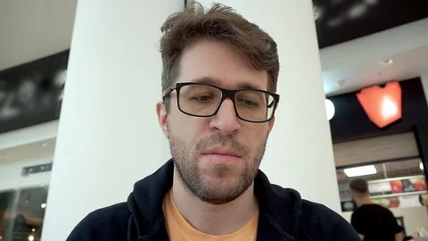 A man in glasses with stubble eats a slice of pizza in a cafe. Видео 100530076