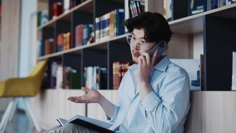 Man in glasses talking on phone while reading book at wooden desk in modern Stock Footage 346892051