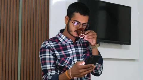 Man with glasses using a smartphone while holding a cup of tea or coffee Видео 329922024