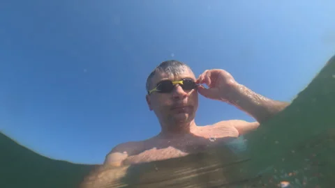 Man with goggles on looking up from below water surface, sun shining through Stock Footage 339822101