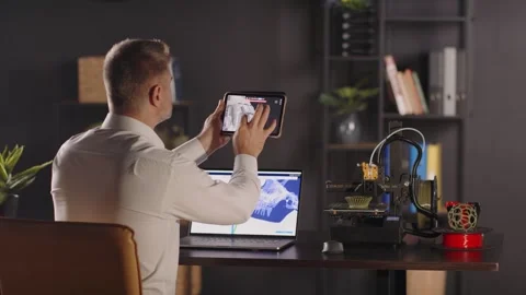 A man graphic designer sits back by the table, uses a tablet to view 3d model Stock Footage 259388737