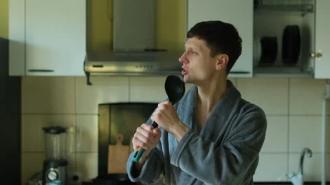 Man in a gray bathrobe playfully singing into a kitchen ladle like a microphone Stock Footage 304632216