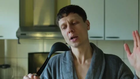 Man in a gray bathrobe singing into a kitchen ladle, enjoying funny everyday Stock Footage 304633389