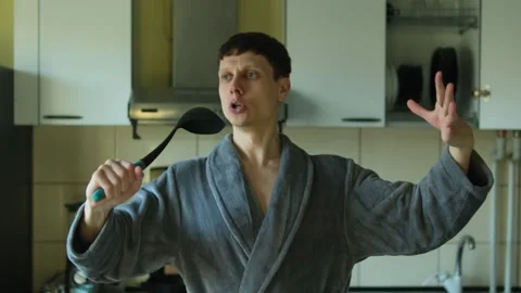 Man in a gray bathrobe singing into a ladle in the kitchen, feeling like a Stock-Footage 304979870
