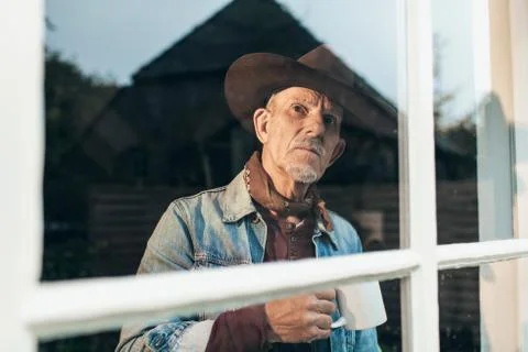 Man with gray beard and hat holding cup of coffee behind window. Stock Photos